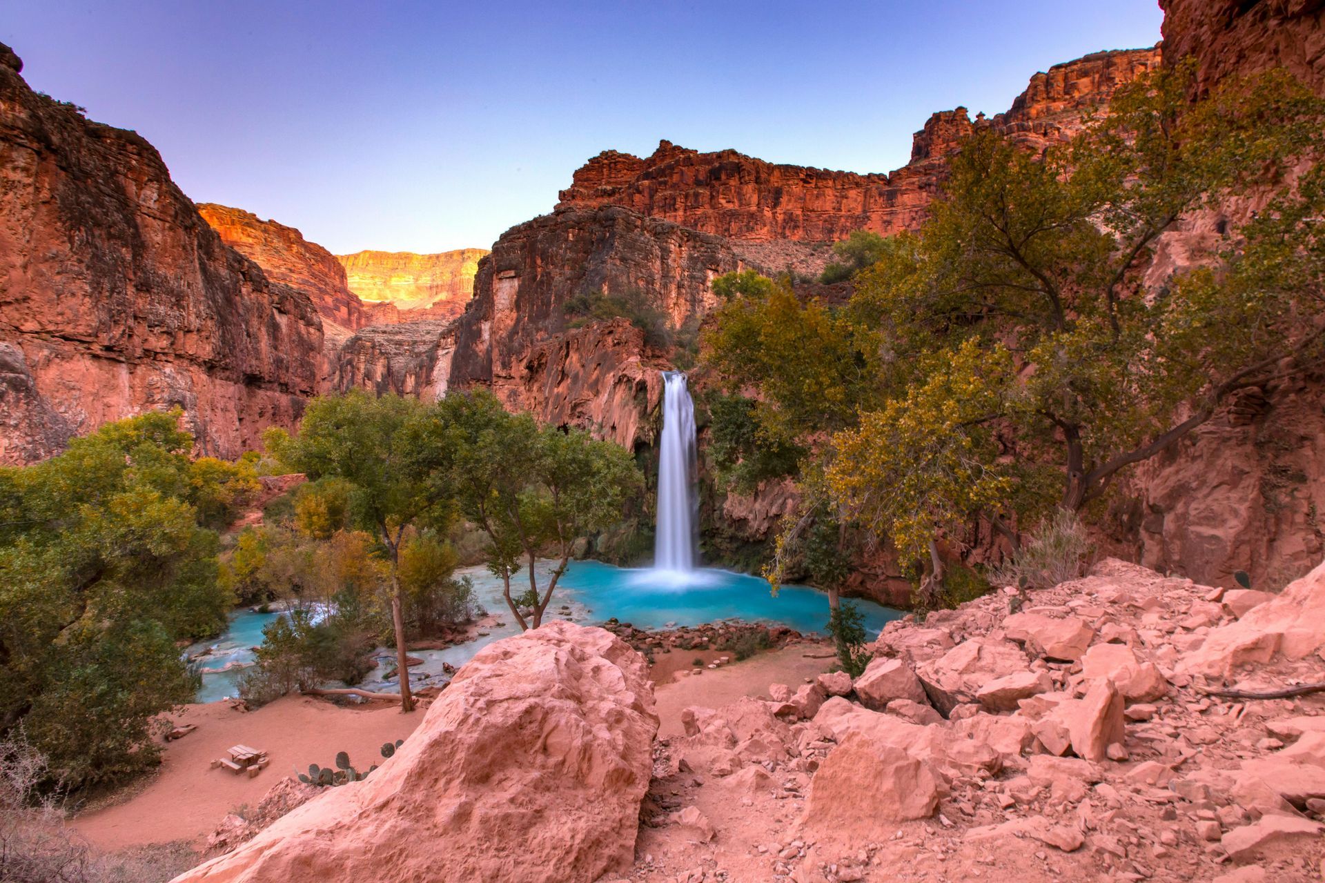 Waterfall cascading into a turquoise pool in a canyon with red rock walls and green trees.
