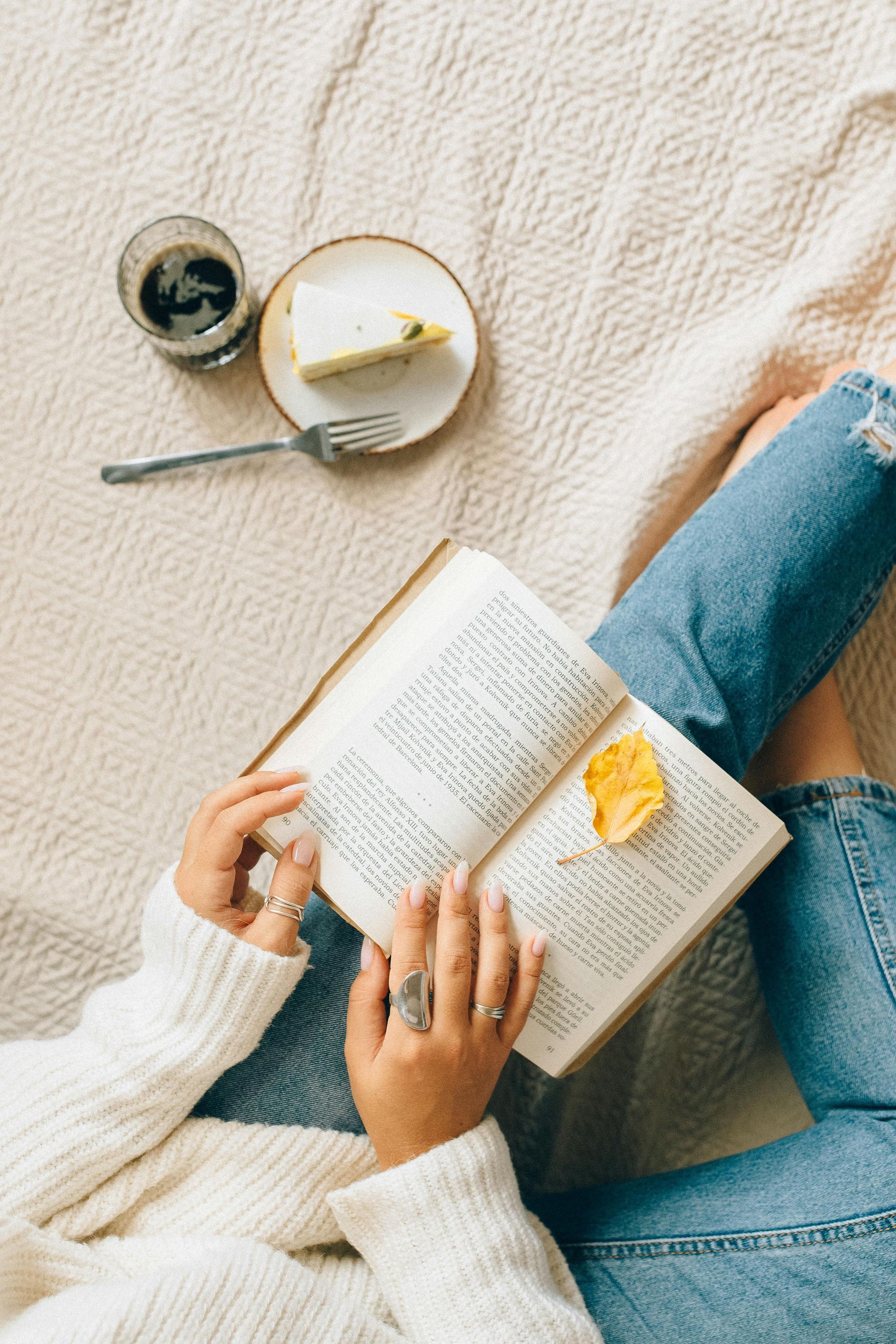 Person in a white sweater and jeans reading a book, with coffee and cake on a bed.