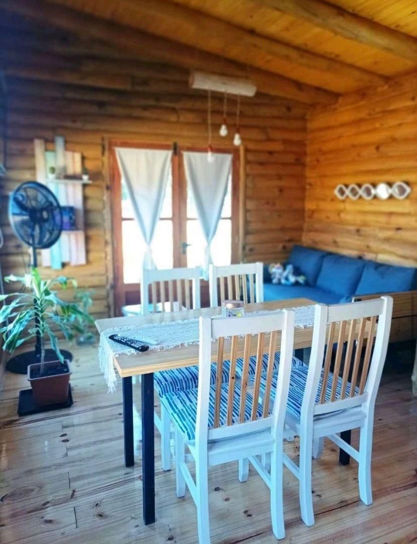 Interior de cabaña de madera con mesa de comedor, sillas y sofá. Paredes y suelos de madera natural; detalles en azul.