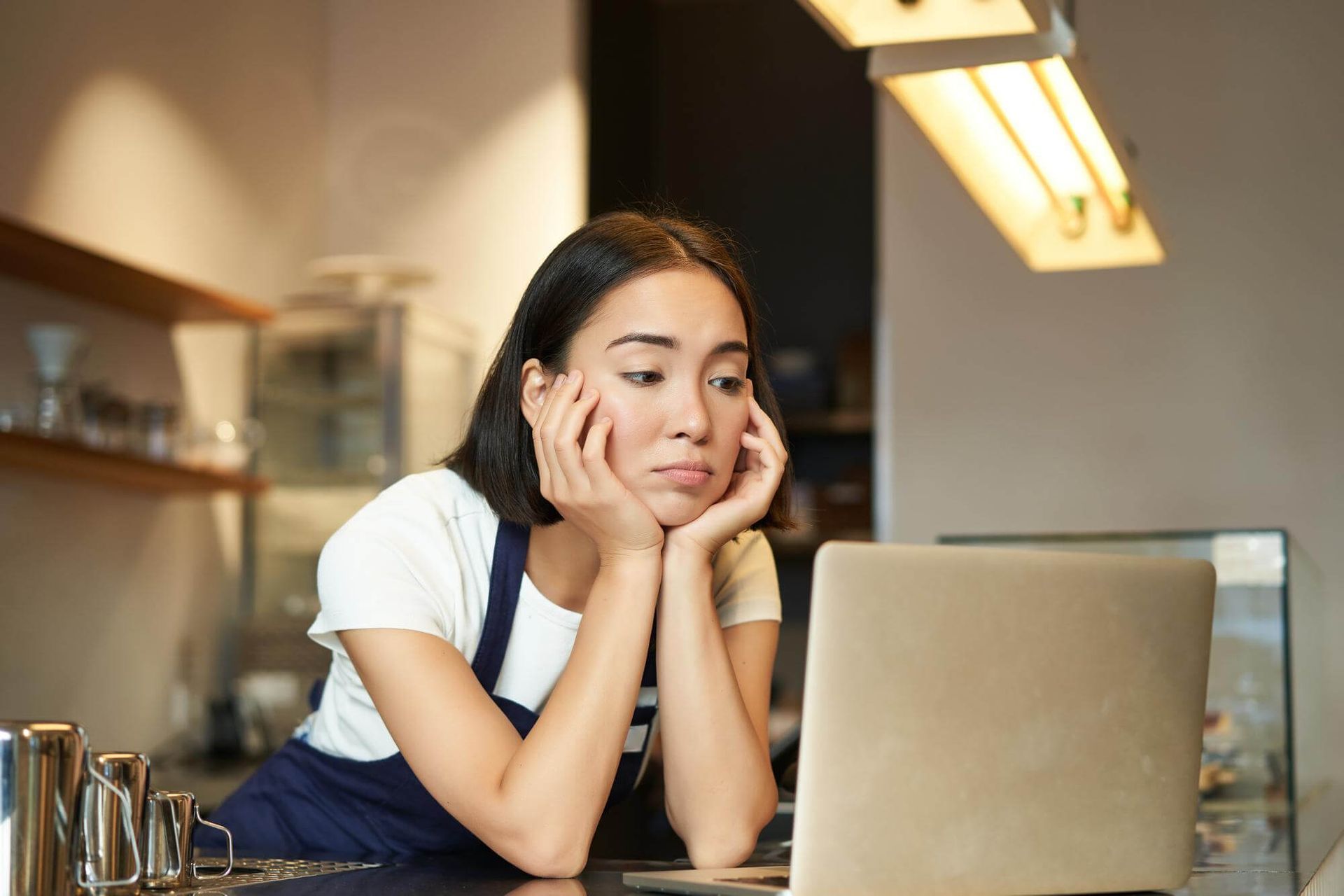 Una mujer con delantal apoya la barbilla en las manos, mirando una computadora portátil en un café.