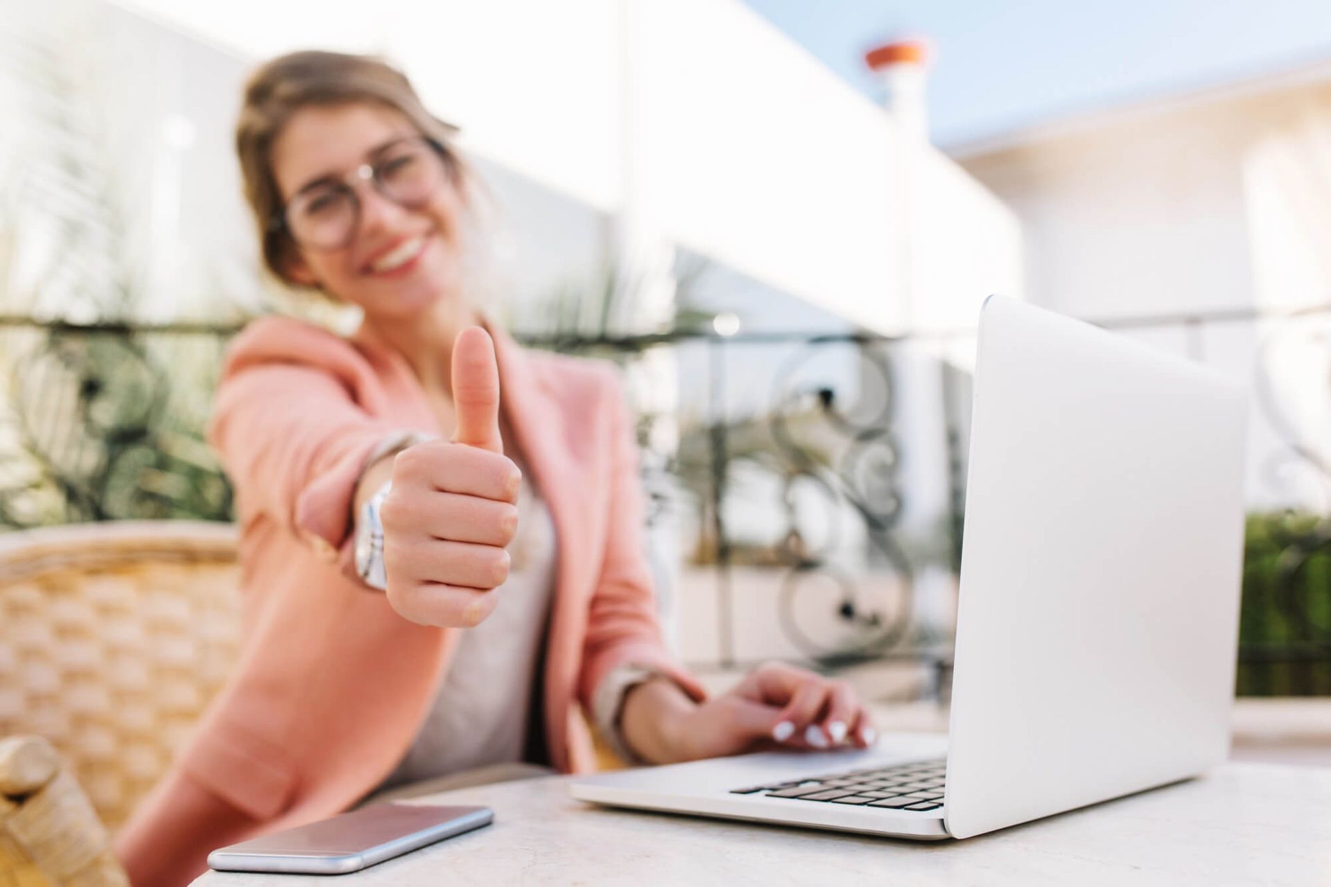 Una mujer con gafas y un blazer rosa levanta el pulgar mientras trabaja con una computadora portátil