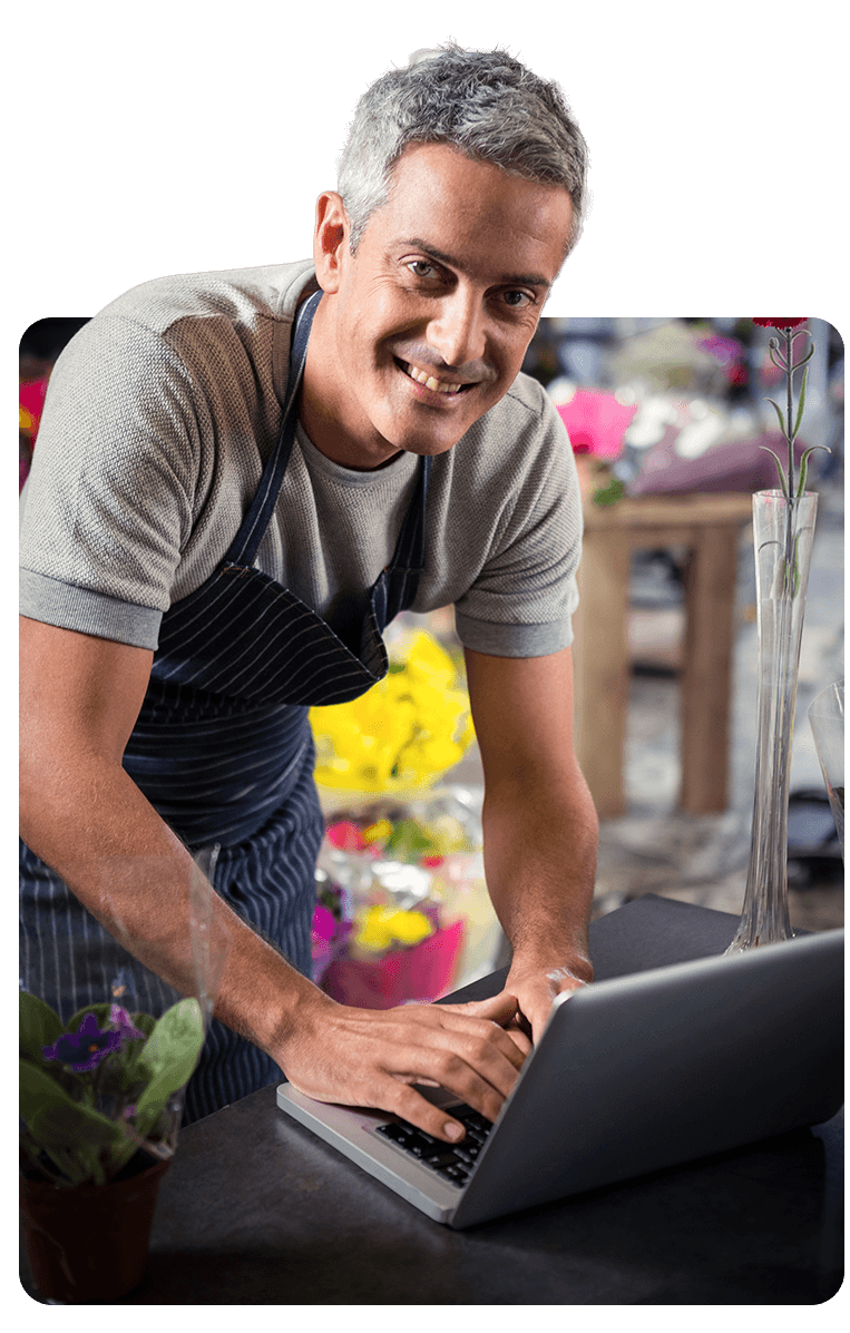 Un hombre con delantal sonríe mientras usa una computadora portátil en una floristería.