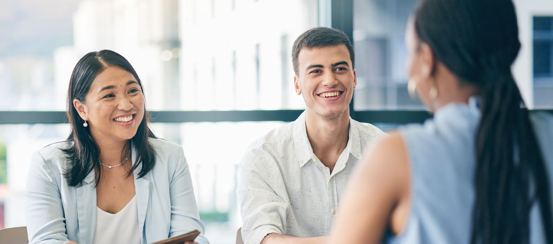 Trois personnes sourient lors d'une réunion. Une femme aux cheveux noirs leur fait face. Un homme et une femme sont assis en face d'elle.