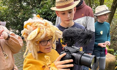 Ein Junge im Löwenkostüm bedient eine Kamera; andere Kinder tragen Hüte und stehen draußen.