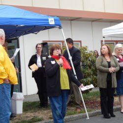 A woman is speaking into a microphone in front of a group of people under a blue tent.