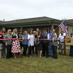 A group of people are standing in front of a house holding a red ribbon.