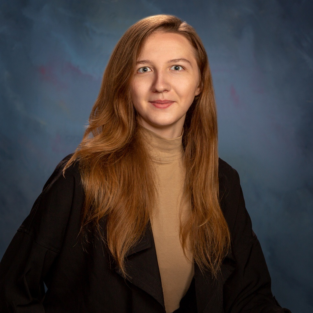 A woman with long brown hair is wearing a black jacket and a tan turtleneck.