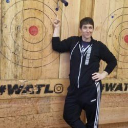 A man in a black jacket is standing in front of a wooden target.