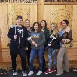 A group of people are standing in front of a wooden target holding axes.