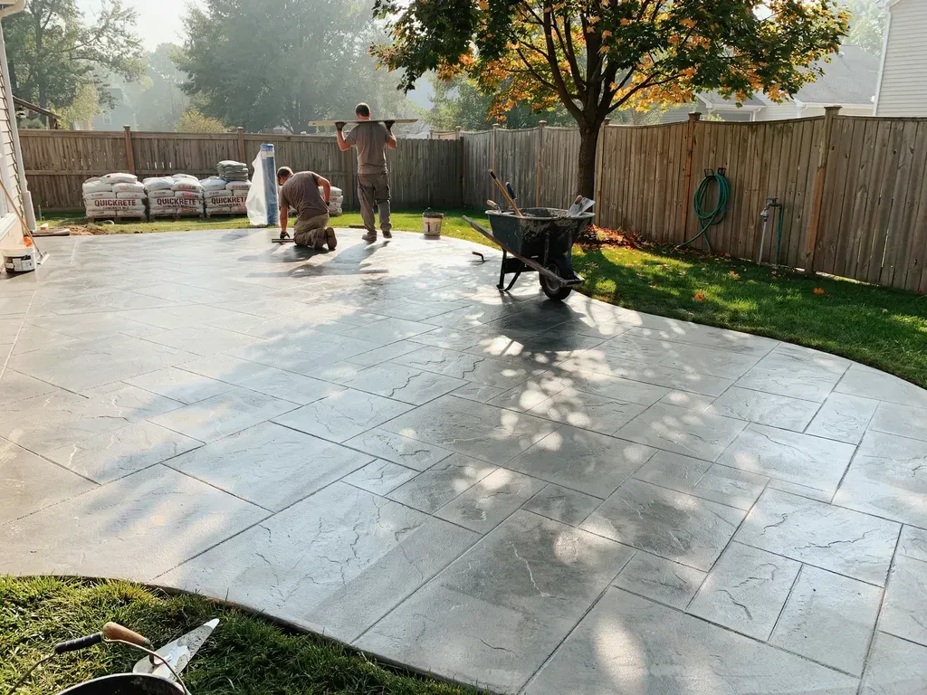 Workers on a sunlit concrete patio near a fence, tree, and wheelbarrow, smoothing the surface