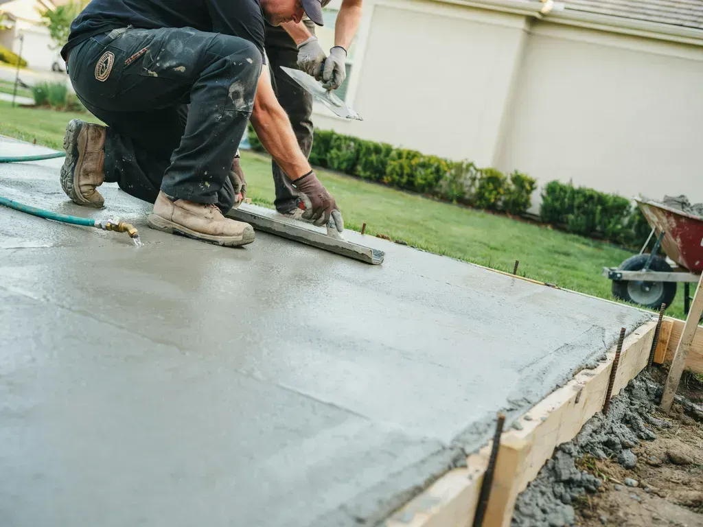 Worker smoothing wet concrete beside a lawn and house foundation during sidewalk or patio construction