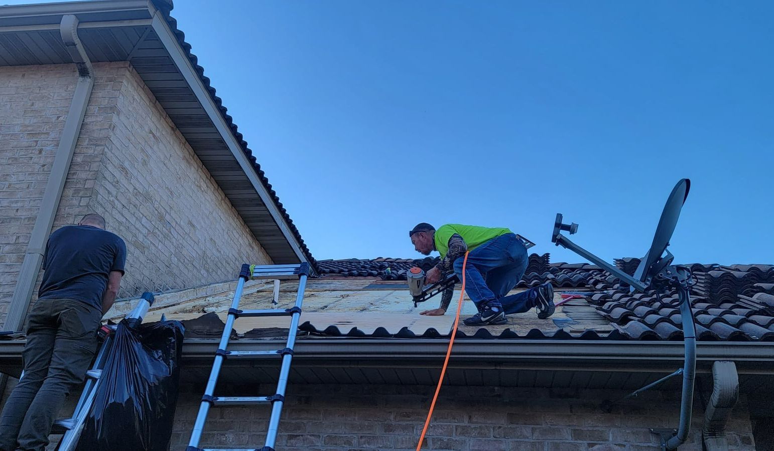 Two workers on a rooftop, one drilling into roofing, ladder present, clear sky.
