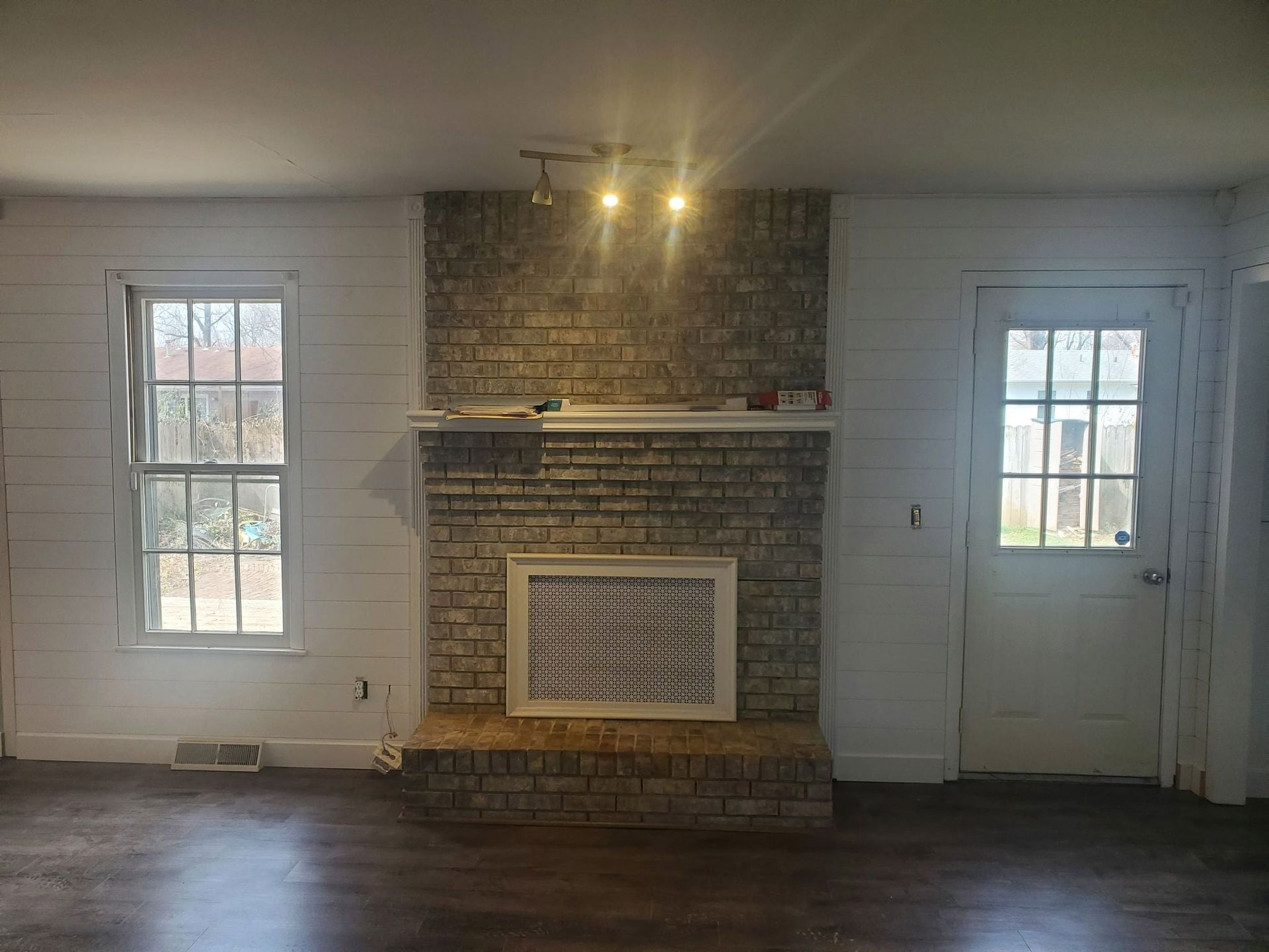 Living room with a brick fireplace between a window and a door, white walls, and wood floor.