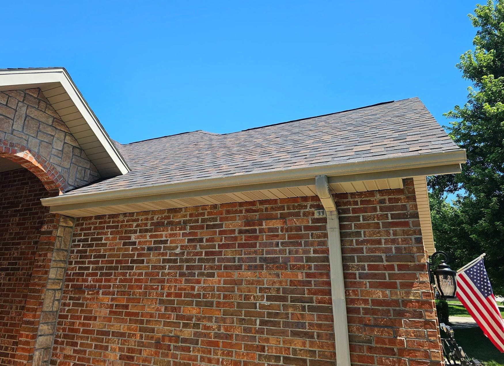Brick house with beige gutters, brown roof, and American flag on a sunny day.
