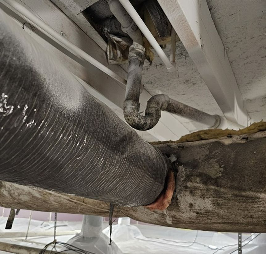 An under-house view with plumbing and a flexible duct covered in white material.