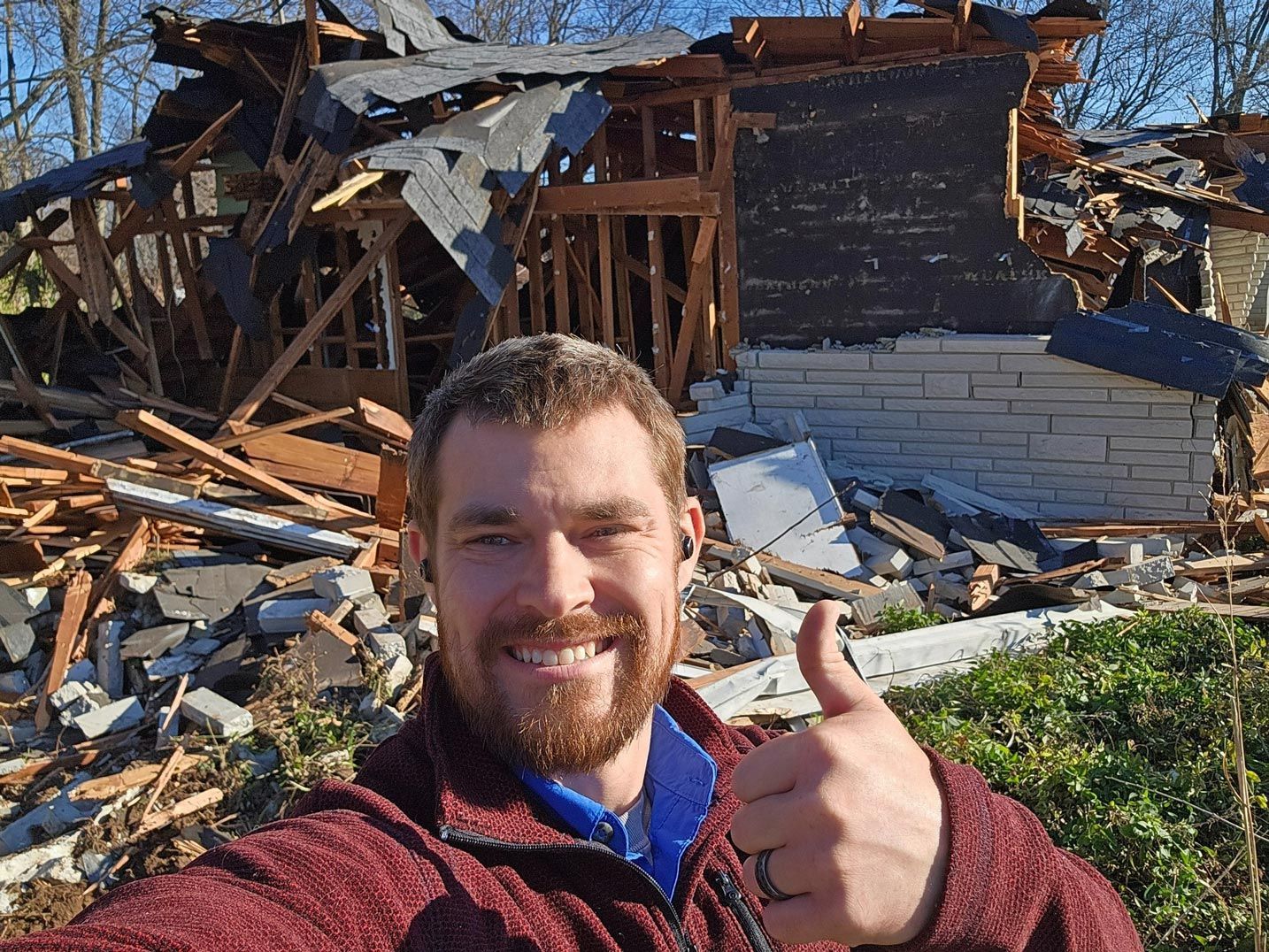 Man smiling, giving thumbs-up in front of a partially demolished house.