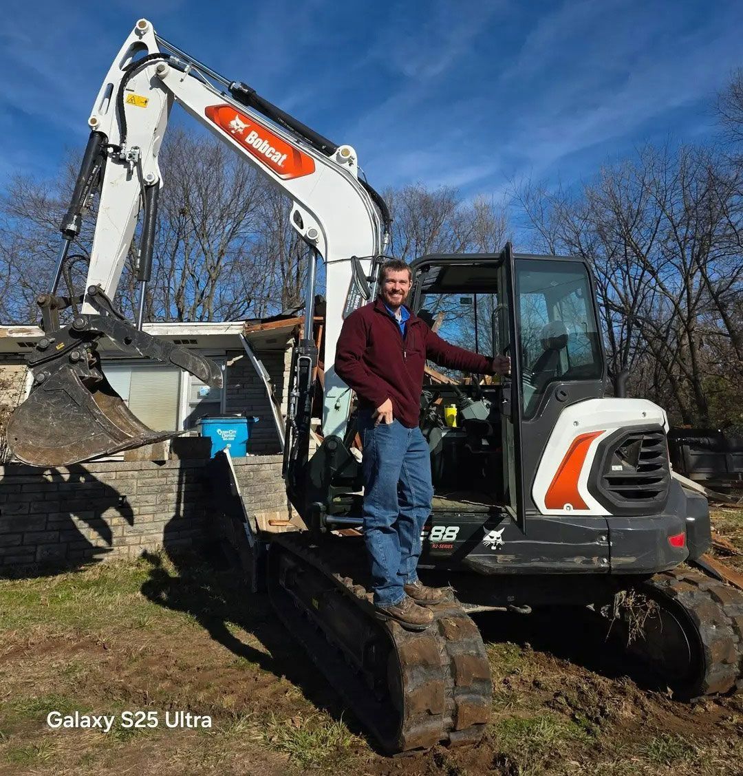 Man in red jacket stands on tracks of Bobcat excavator. Bright blue sky.