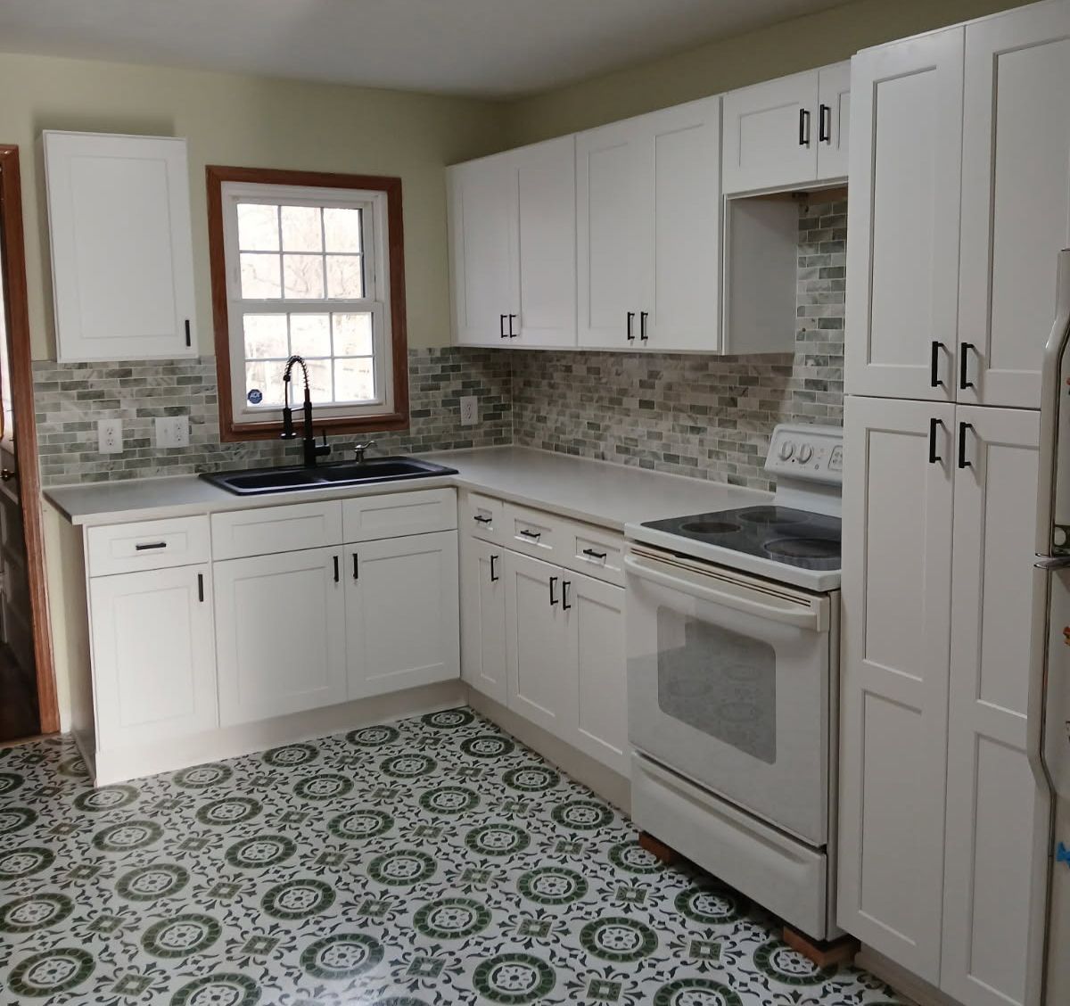 White kitchen cabinets, countertop, stove, and patterned floor. Window with black faucet.