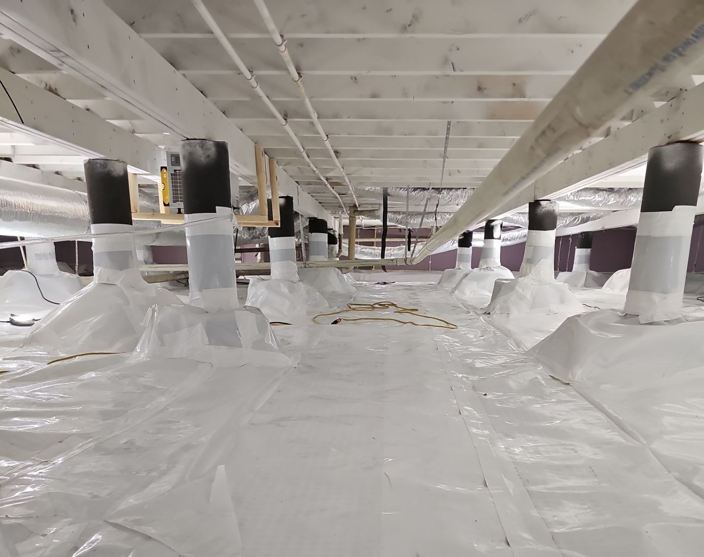 Crawl space with white vapor barrier, support posts, and wooden joists.
