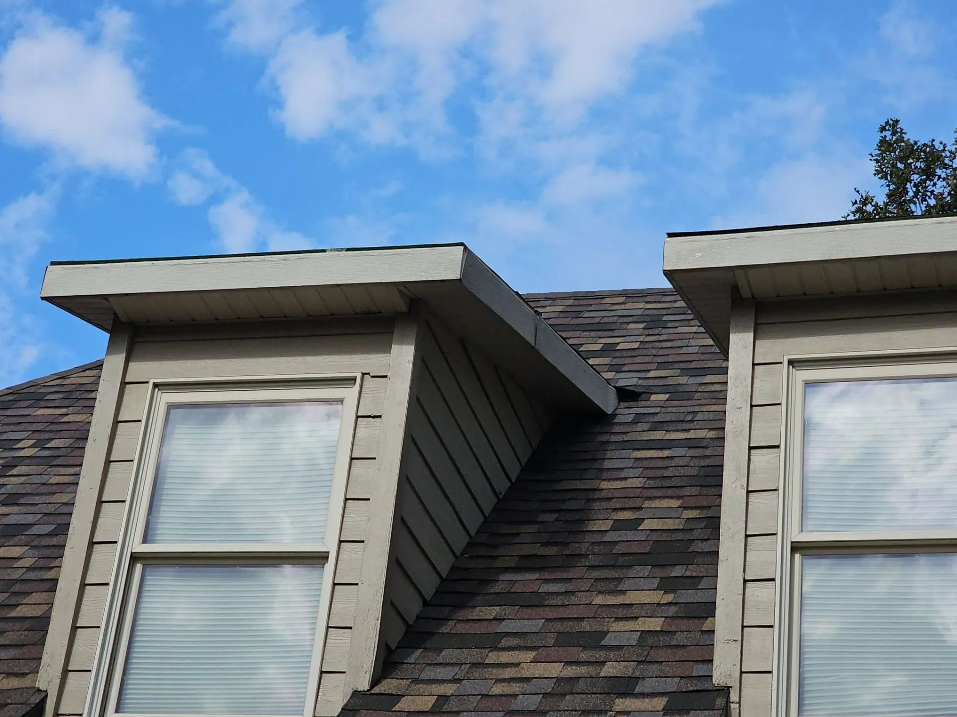 Two dormer windows on a roof with asphalt shingles against a blue sky with clouds.