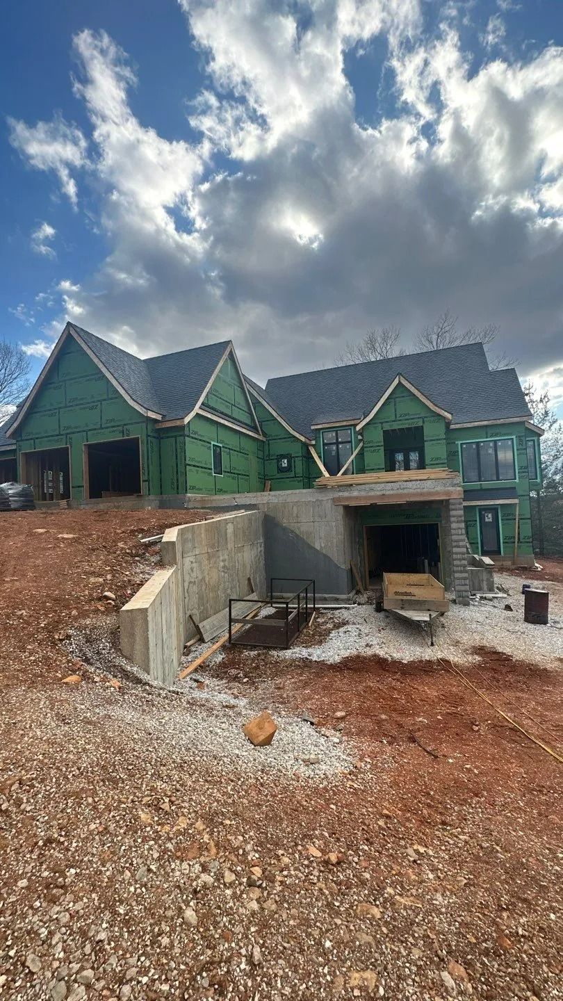 House under construction with green wrap, unfinished garage, and overcast sky.