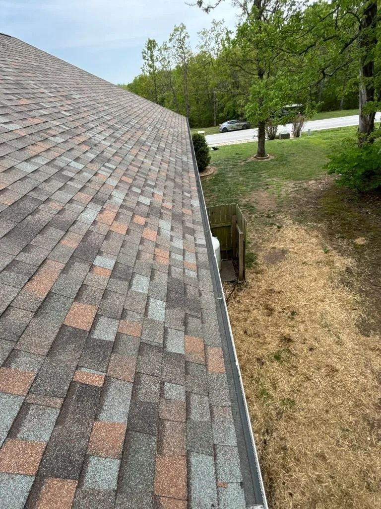 Roof shingles with shades of brown, gray, and red. Gutter along the edge, trees, and dry grass.