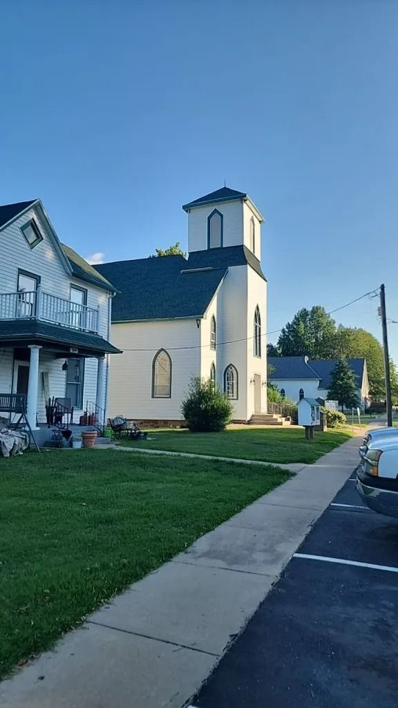 White church building with a tower next to a sidewalk and houses on a sunny day.