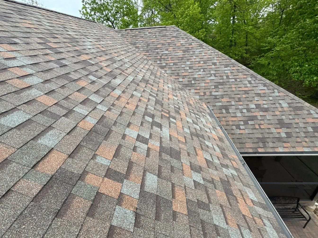 Shingled roof with various shades of brown, gray, and orange. Lush green trees in background.