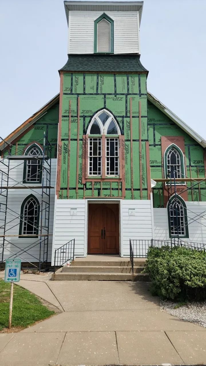 Church under renovation, green sheathing, white siding, arched windows, and scaffolding.