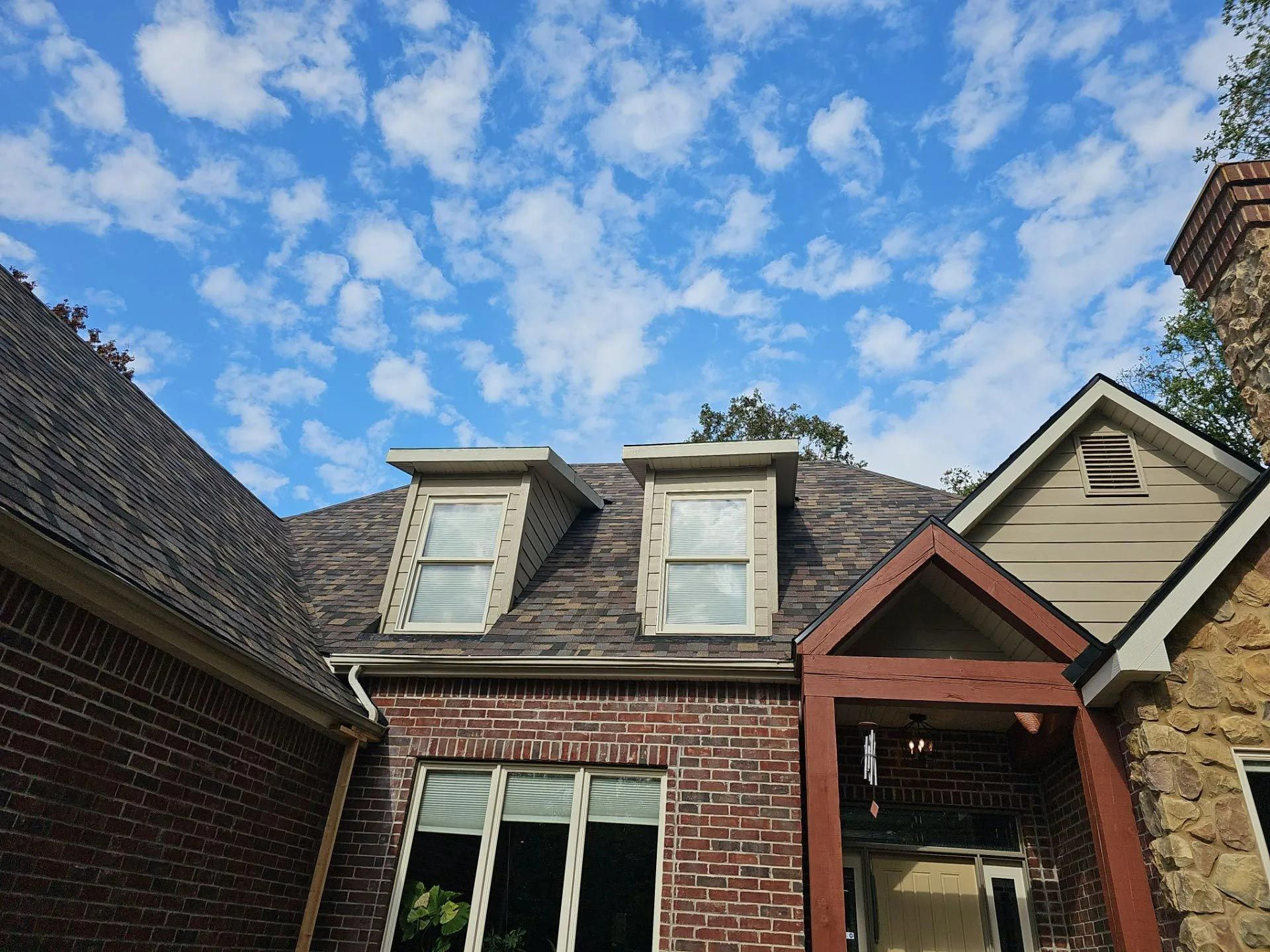 Brick house with brown roof, dormer windows, and a blue sky dotted with clouds.