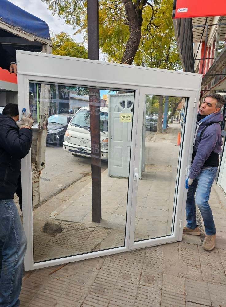 Dos hombres llevan un gran marco de ventana de vidrio con borde blanco en una acera frente a una tienda.