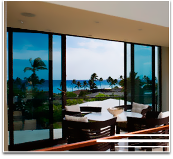 Balcón con vista al mar a través de puertas corredizas de vidrio tintado azul. Muebles blancos en la terraza. Palmeras.