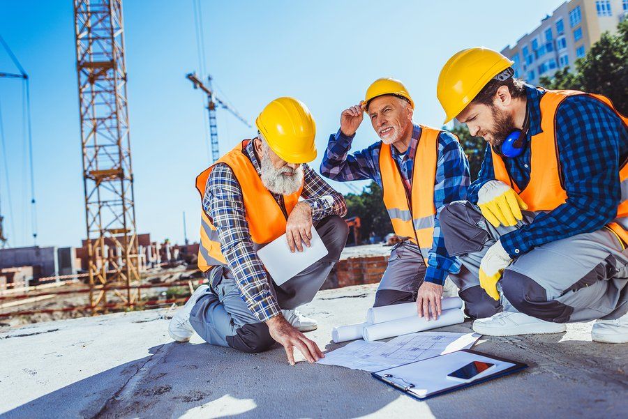 Three construction workers in uniform sitting on concrete at construction site examining building plans