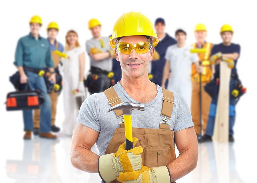 Group of construction workings lined up with their yellow hard hats on in the background and a man standing in the front with a hammer, gloves and hard hat on.