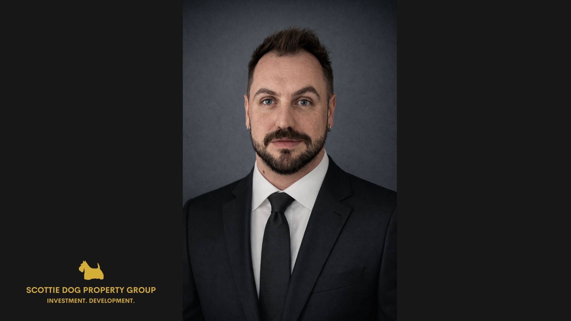 A man in a suit and tie poses against a grey backdrop, with a small gold logo in the bottom-left corner.