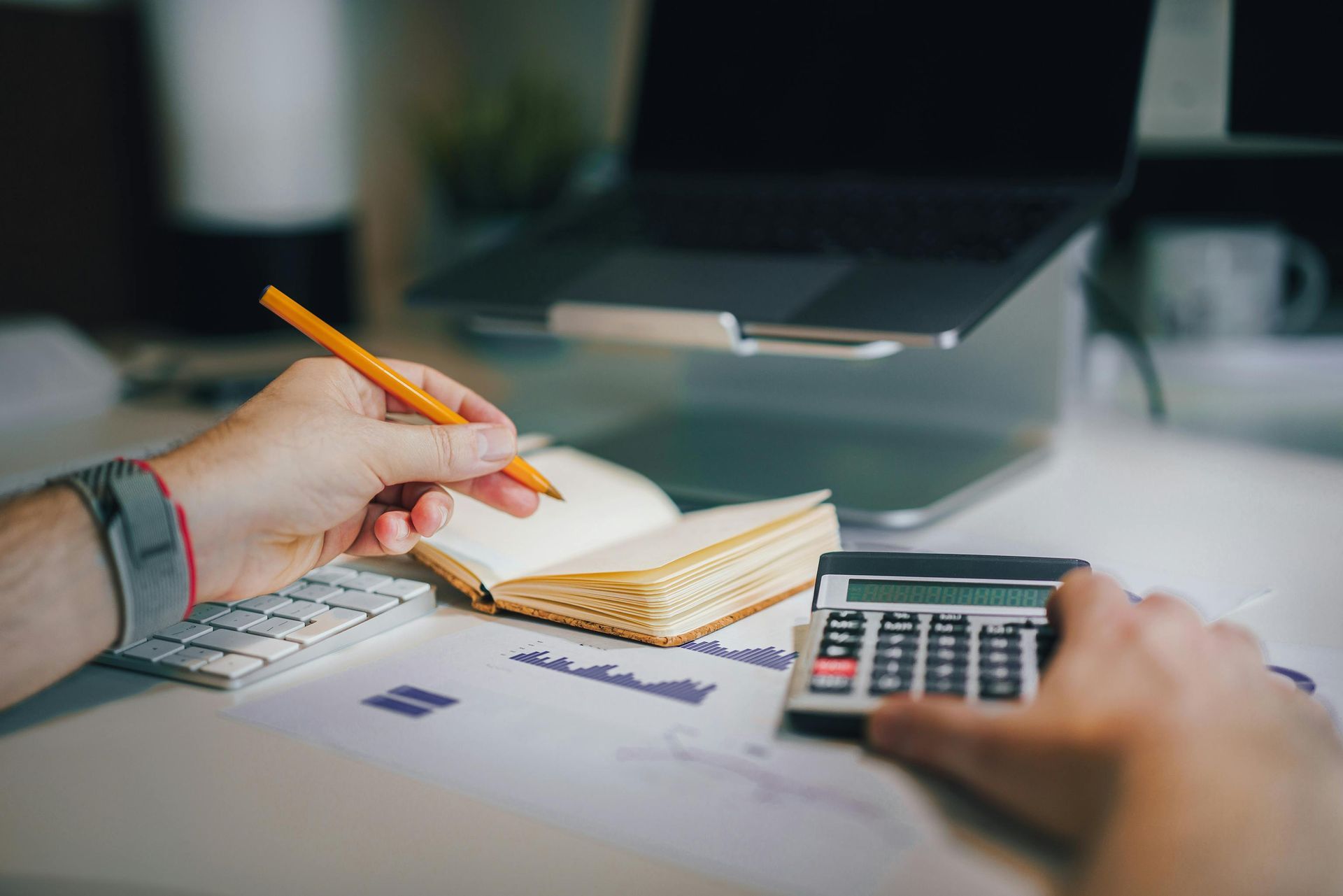 Hands at a desk using a calculator, pen, and notebook next to a laptop on a stand.