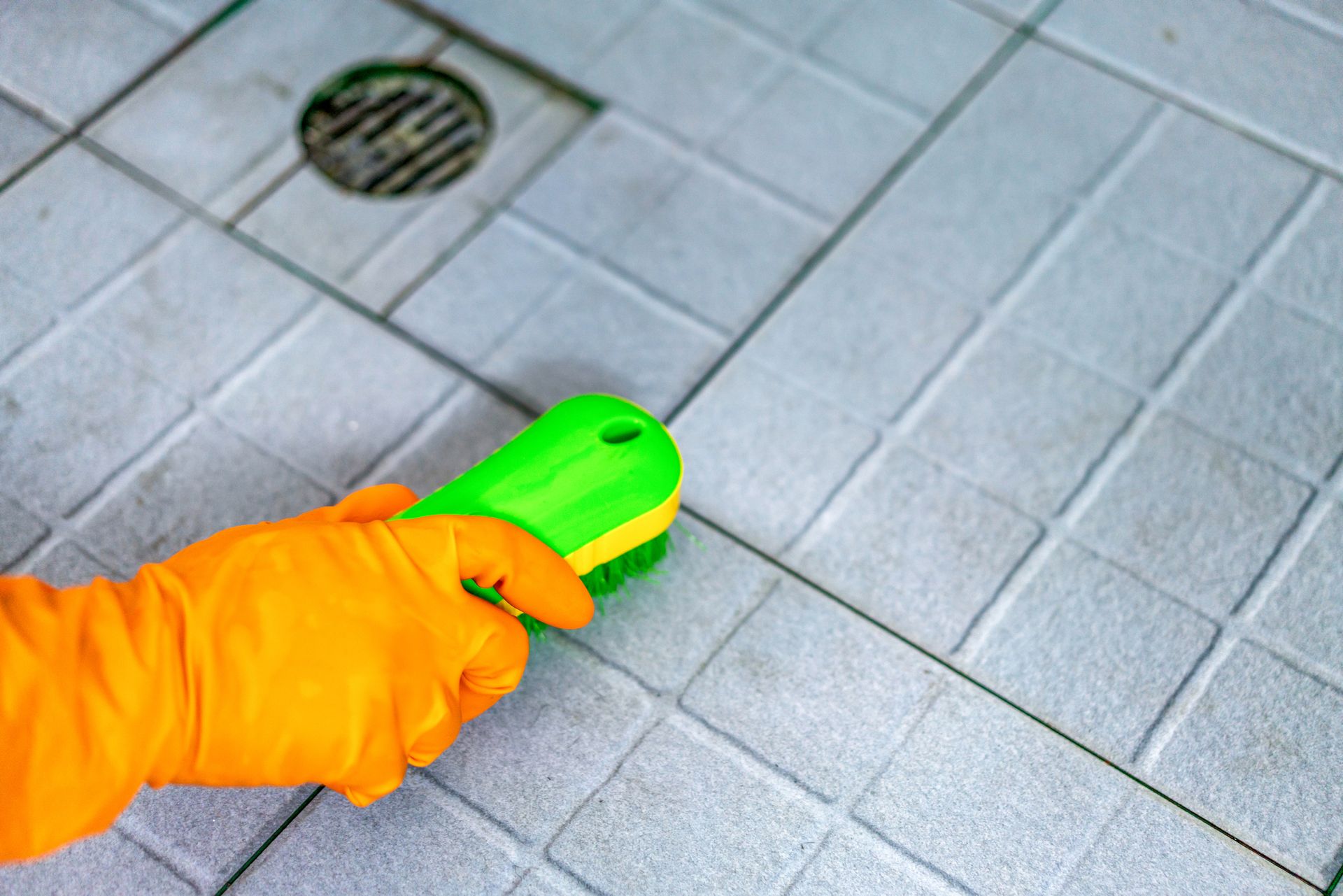 A person wearing orange gloves is cleaning a tile floor with a green brush.