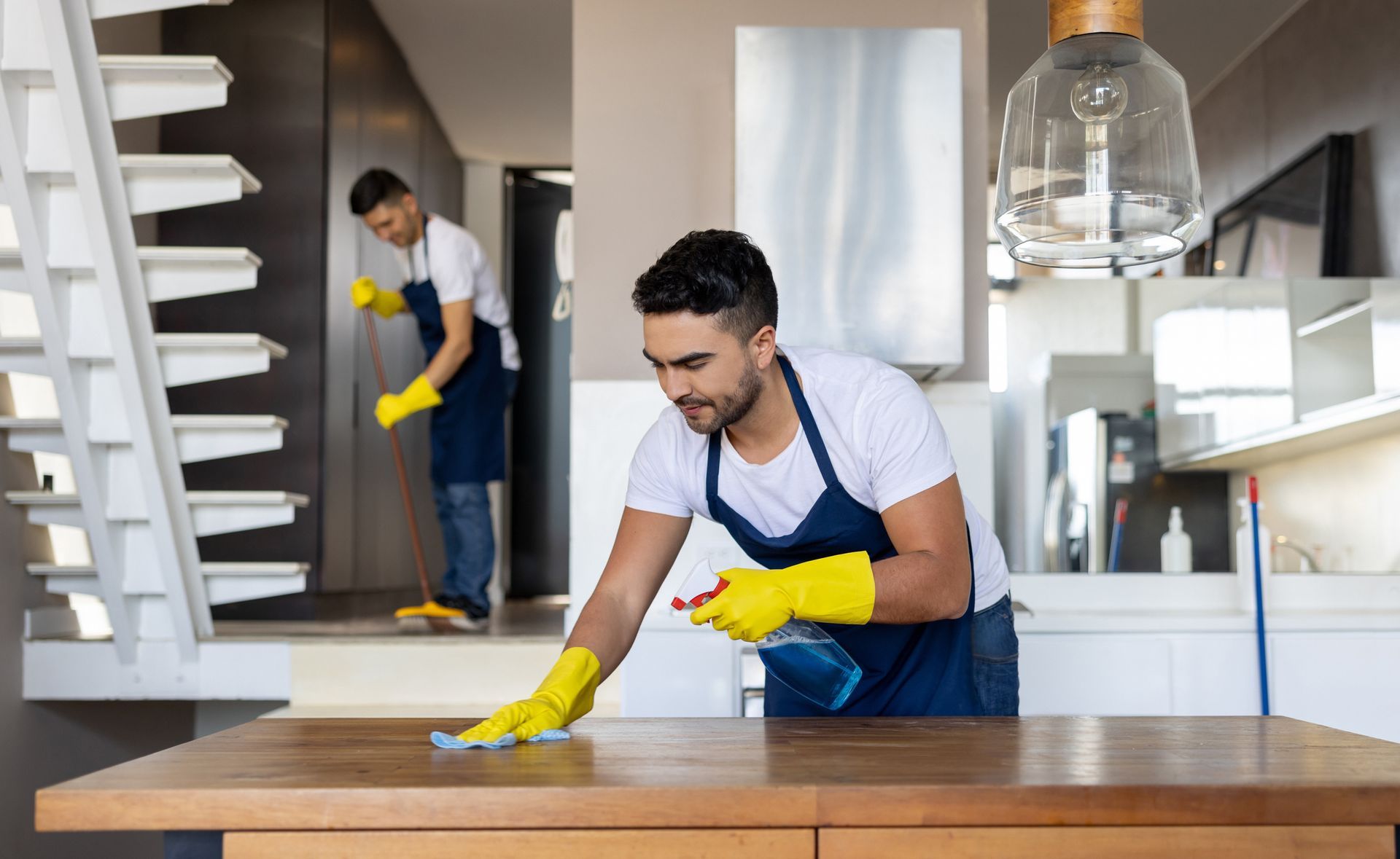 Two men are cleaning a wooden table in a kitchen.