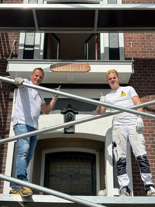 Twee schilders op een steiger steken hun duim omhoog voor de ingang van een huis.