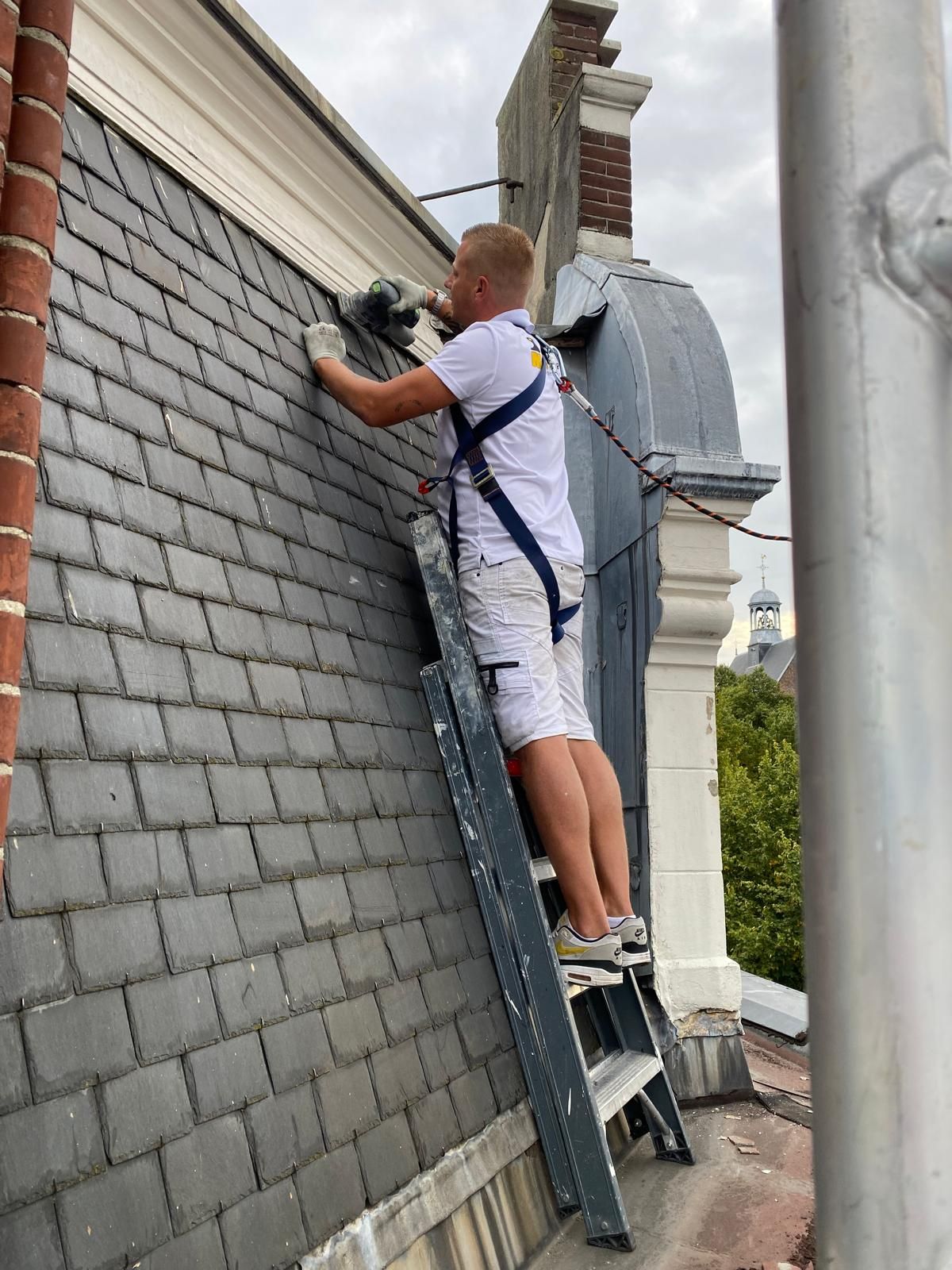 Man op een ladder repareert een leien dak. Hij draagt ​​een veiligheidsharnas en werkt met gereedschap, bewolkte lucht.