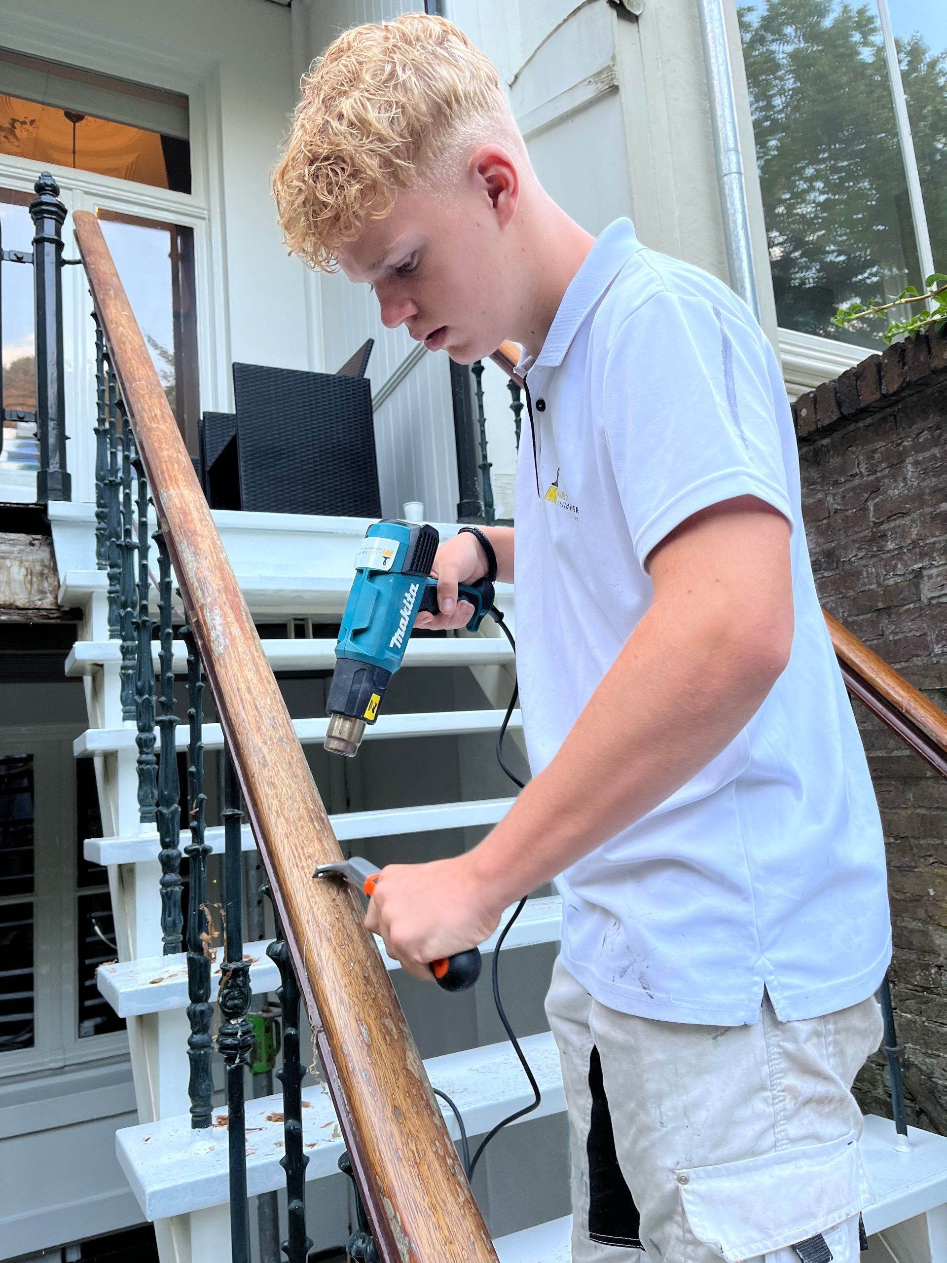 Persoon die een boormachine en een klein handgereedschap gebruikt op een houten trapleuning buitenshuis.