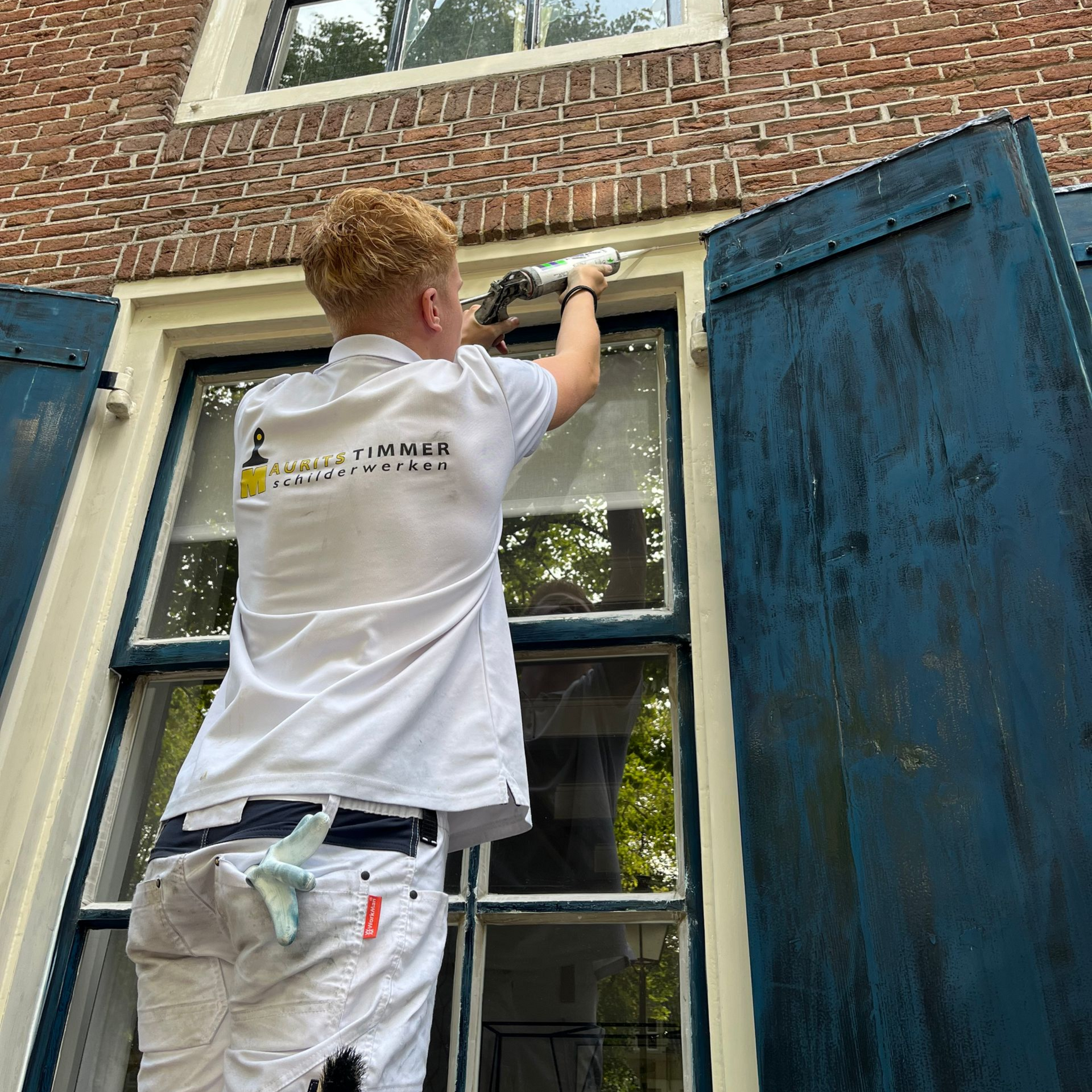 Persoon die het kozijn van een bakstenen gebouw met blauwe luiken afkit.