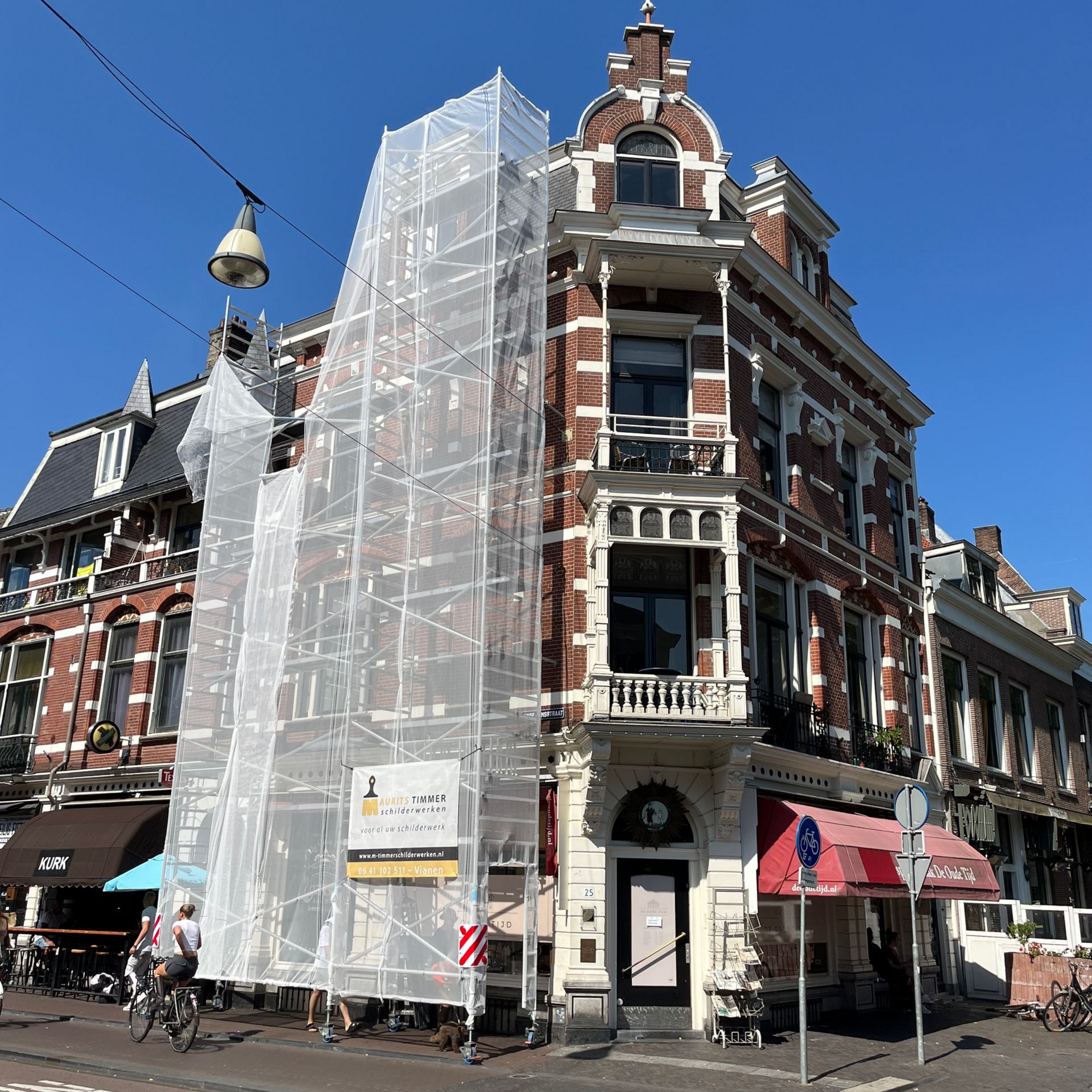 Hoek van het gebouw met steigerwerk en wit net. Straatniveau met fietsen, luifel en een zonnige hemel.