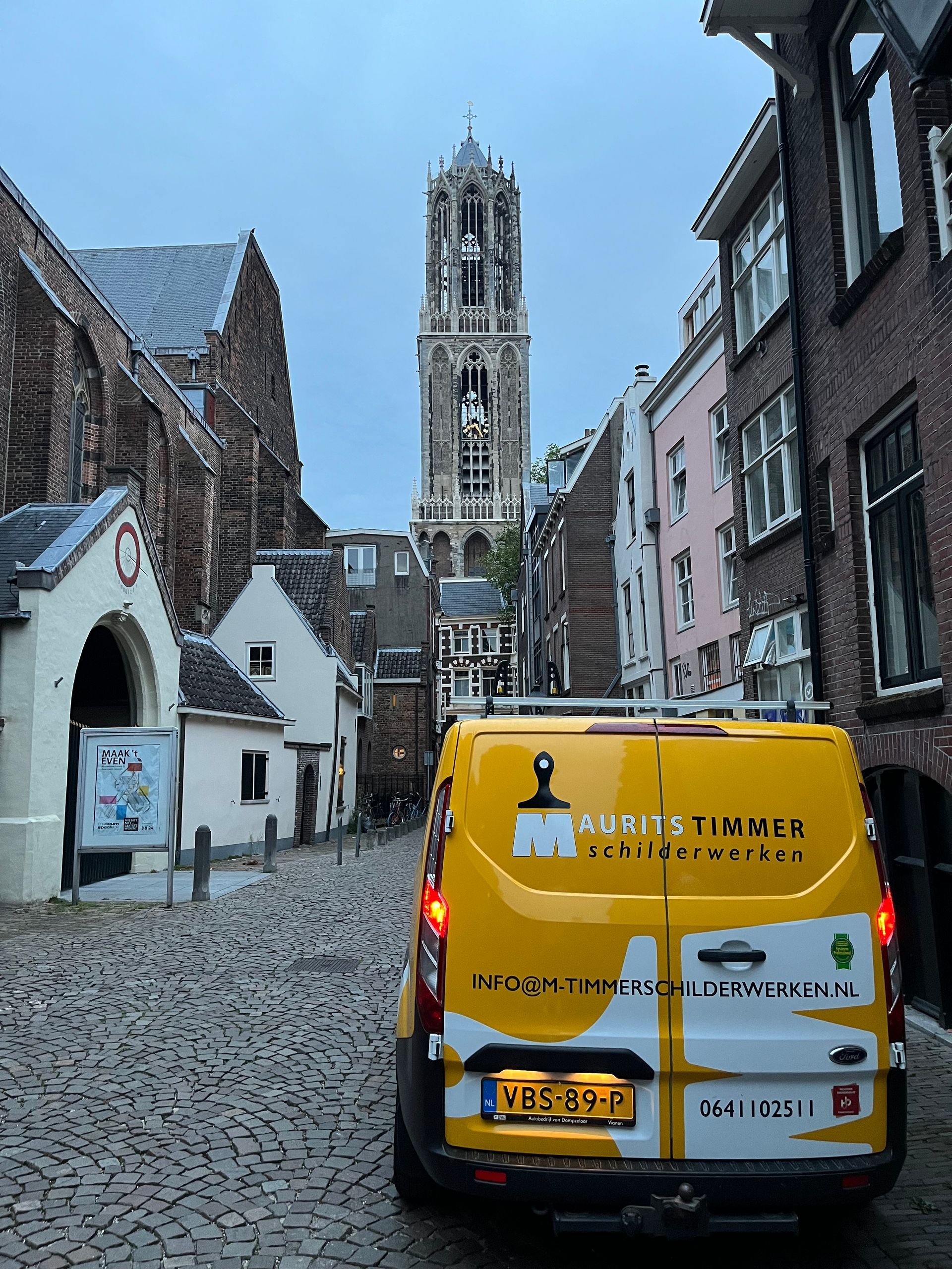 Geel busje geparkeerd op de geplaveide straat, historische gebouwen, hoge kerktoren op de achtergrond.
