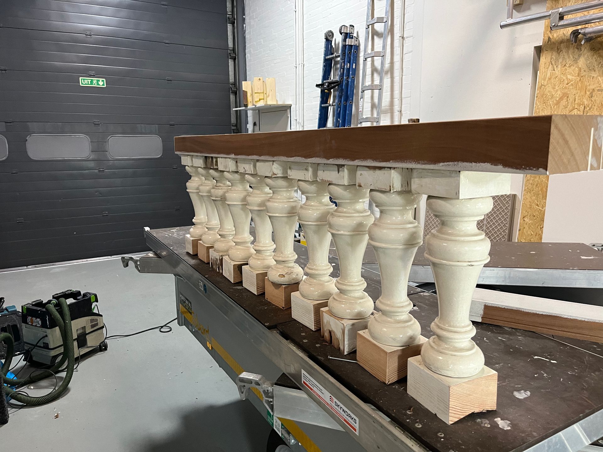 Een gedeeltelijk gemonteerde balustrade van witte houten spijlen op een werkbank, met een donkerbruine bovenregel.