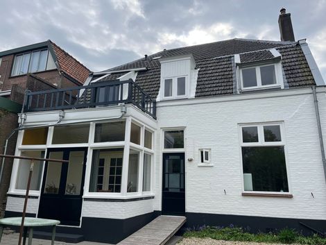 Wit bakstenen gebouw met donkere randen, balkon en helling die naar een zwarte deur leidt, bewolkte lucht.