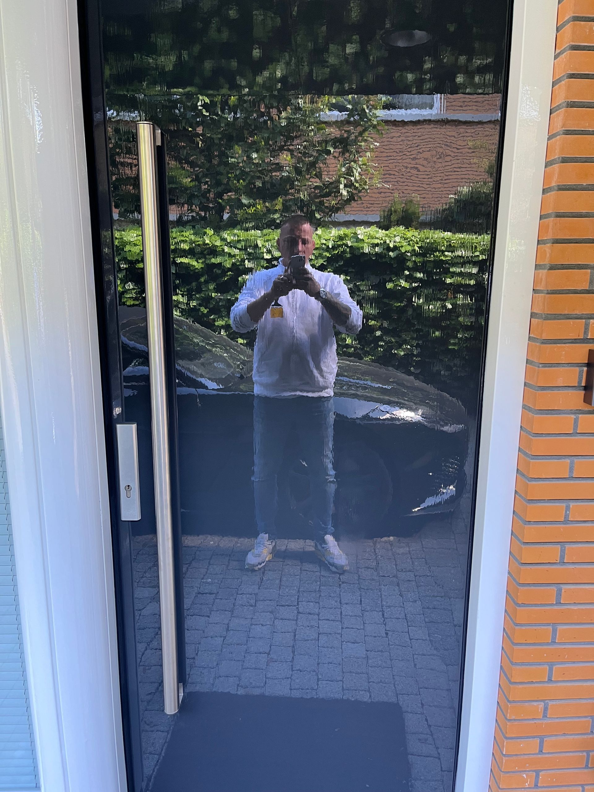 Man maakt een foto door een zwarte glazen deur waarin zijn omgeving wordt weerspiegeld: een auto, bomen en een bakstenen huis.