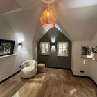 Gezellige zolderkamer met witte muren, een houten vloer en een decoratieve geweven hanglamp. Inclusief een stoel, een voetenbank en twee ramen.