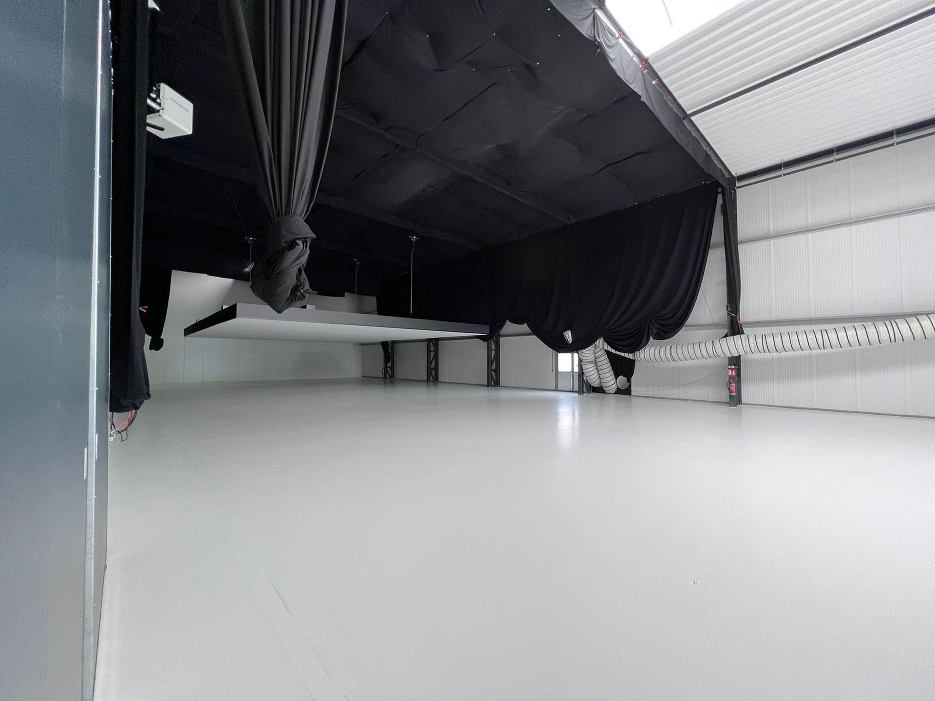 Lege kamer met een witte vloer, zwarte gordijnen aan het plafond, metalen wanden en een zichtbare steunbalk.