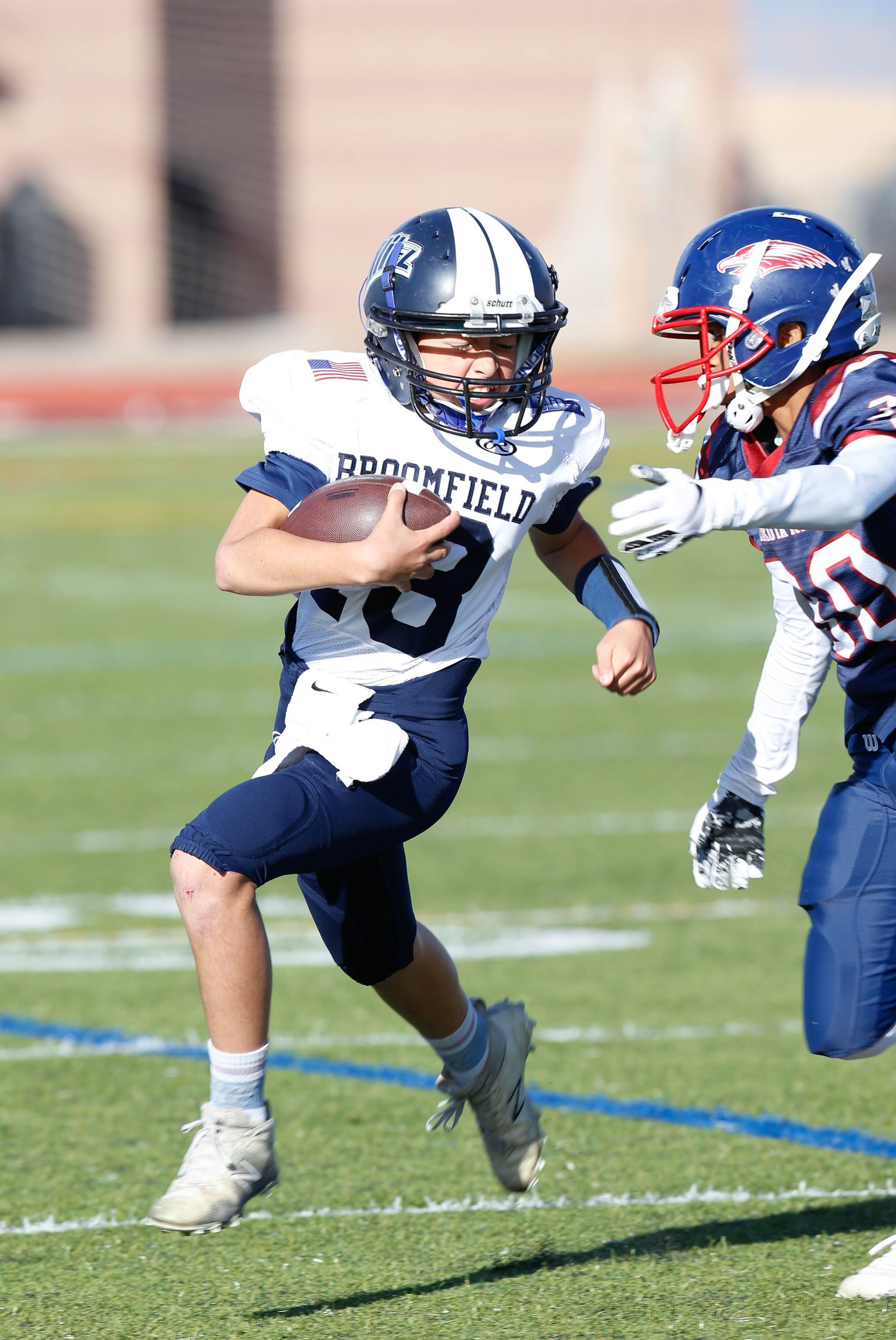 Football players in action on a grassy field; one runs with the ball, pursued by two opponents.